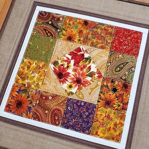Peut inclure: Un carré de courtepointe en tissu présentant un motif central en losange à fleurs, entouré de divers carrés en patchwork aux couleurs automnales comme l'orange, le rouge, le vert et le violet, le tout encadré par une large bordure beige et marron.