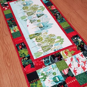 May include: A rectangular quilted table runner featuring a central panel with a stylised winter forest scene in shades of green, blue, and white. The border consists of a patchwork of squares in red, green, black, and white with floral and bird motifs.