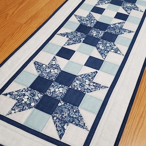May include: A rectangular quilted table runner featuring a star pattern. The stars are made of floral fabric in shades of blue and white, set against a light blue and navy blue checkerboard design. The runner is bordered with a light gray fabric.