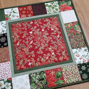 May include: A square quilted table mat featuring a central red panel with a gold-outlined poinsettia and berry pattern, surrounded by a border of patchwork squares in red, green, white, and black with various holiday-themed floral prints.
