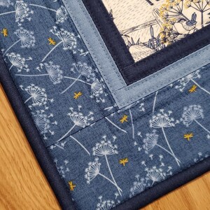 May include: A quilted placemat with a blue dandelion pattern and a central panel featuring a deer and bird scene. The placemat has a dark blue border and light blue accent strips.