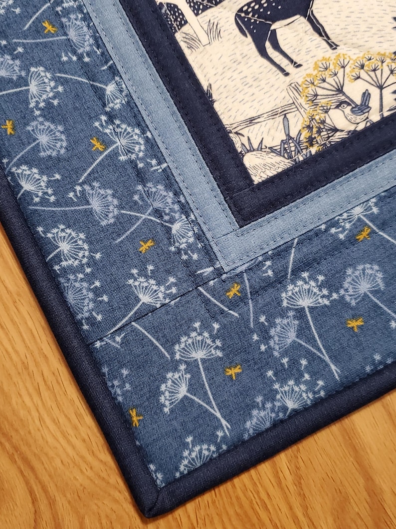 May include: A quilted placemat with a blue dandelion pattern and a central panel featuring a deer and bird scene. The placemat has a dark blue border and light blue accent strips.