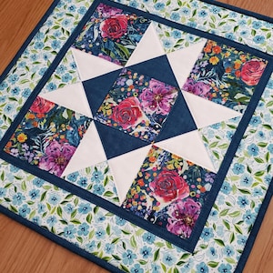 Puede incluir: Camino de mesa acolchado con un estampado floral. El centro presenta un diseño de estrella con triángulos blancos y cuadrados florales en tonos de rosa, morado y azul. El borde tiene un estampado floral azul con un ribete azul oscuro.
