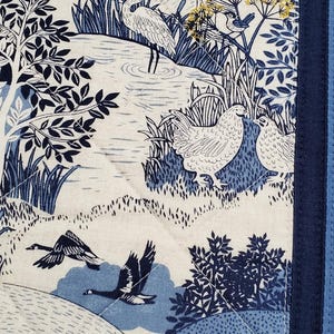 May include: A quilted fabric panel featuring a pastoral scene in shades of blue and white. The design includes illustrations of birds, geese, trees, and a body of water. The panel is bordered with a dark blue fabric and has a quilted pattern.