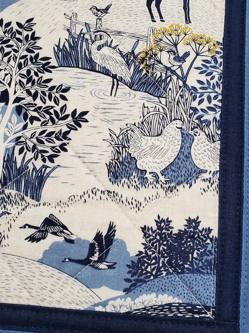 May include: A quilted fabric panel featuring a pastoral scene in shades of blue and white. The design includes illustrations of birds, geese, trees, and a body of water. The panel is bordered with a dark blue fabric and has a quilted pattern.