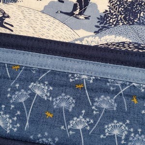 May include: A quilted fabric with a nature-themed design. The top section features a cream-colored background with blue geese and a rabbit. The bottom section is blue with white dandelion-like flowers and yellow dragonfly accents.