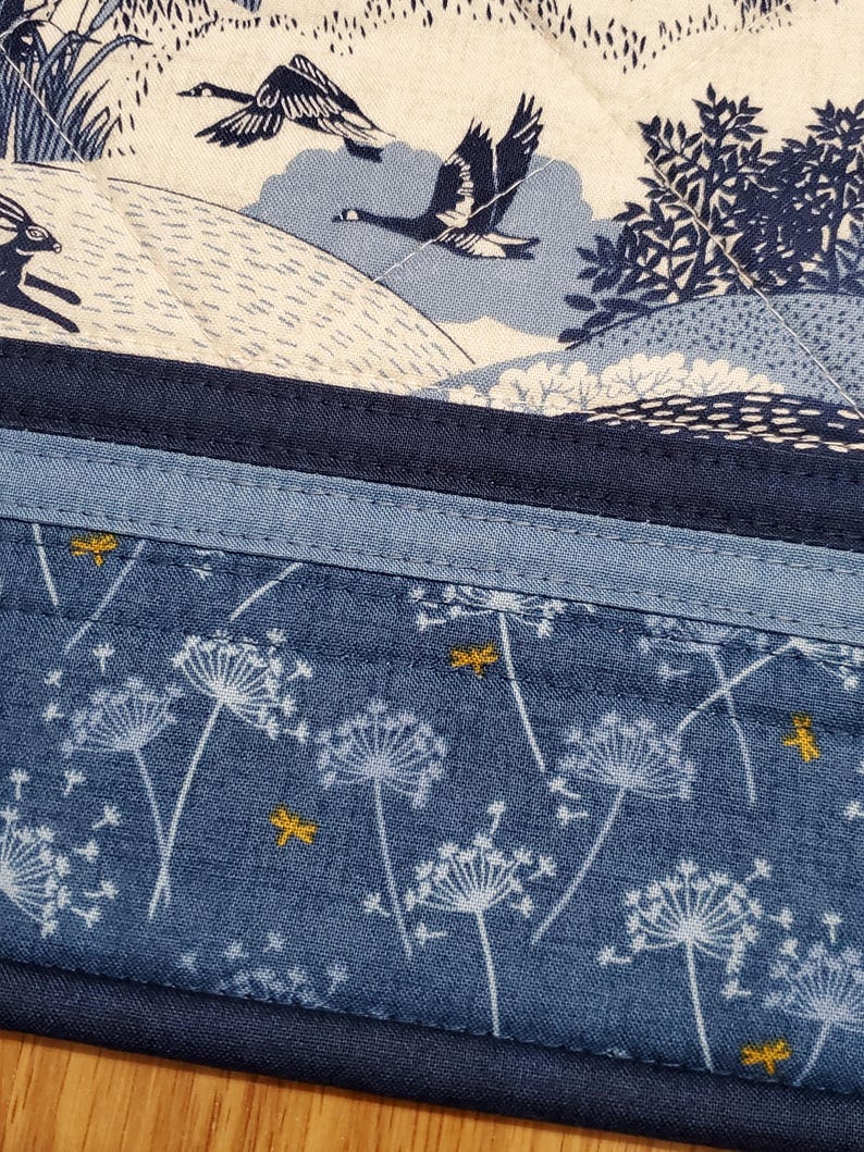 May include: A quilted fabric with a nature-themed design. The top section features a cream-colored background with blue geese and a rabbit. The bottom section is blue with white dandelion-like flowers and yellow dragonfly accents.