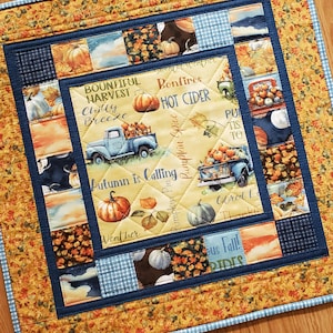 May include: A square quilted fabric wall hanging featuring a central panel with pumpkins, a blue truck, and text including Bountiful Harvest, Hot Cider, and Autumn is Calling. The border consists of patchwork squares in autumn colours and blue.