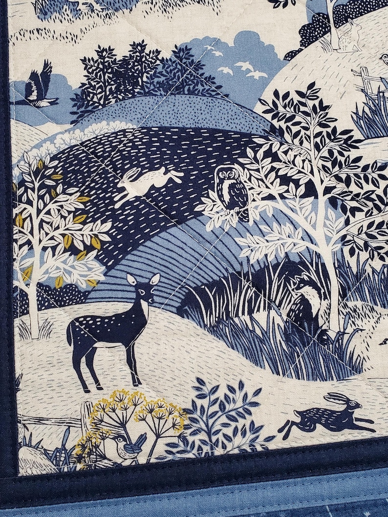 May include: A quilted fabric with a nature scene in shades of blue and white. The design features a deer, rabbits, an owl, trees, and birds. The fabric is bordered with a dark blue trim.