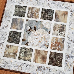 May include: A square fabric quilt featuring a central panel with a large white peony and cotton boll print. The center is surrounded by twelve smaller square patches in shades of brown, beige, and gray, framed by a white border and floral edge.