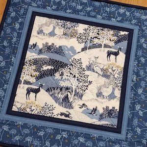 May include: A quilted placemat with a nature scene. The center features a detailed illustration of deer, rabbits, birds, and trees in shades of blue and white. The placemat is bordered by a blue floral pattern and a solid navy blue binding.