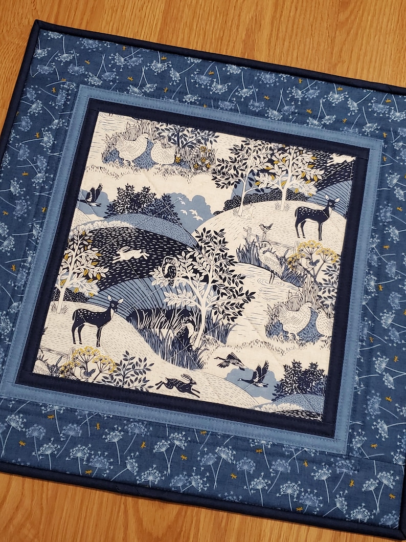 May include: A quilted placemat with a nature scene. The center features a detailed illustration of deer, rabbits, birds, and trees in shades of blue and white. The placemat is bordered by a blue floral pattern and a solid navy blue binding.