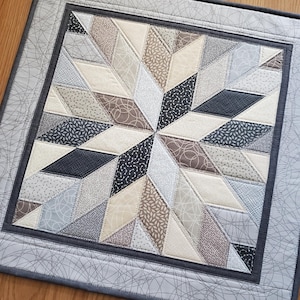 May include: A handcrafted quilt featuring a central starburst design. The star is composed of various fabric patterns in neutral tones of beige, gray, and black. The quilt is framed with a gray border and a decorative stitch pattern.