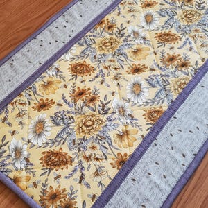 May include: A quilted table runner with a floral pattern in yellow, brown, and white. The runner has a lavender border with text and bee illustrations. The center features a repeating pattern of flowers and bees.