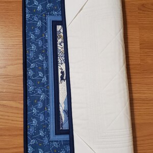 May include: A partially rolled-up quilted placemat with a dark blue border. The front features a blue floral pattern and a central panel with a rabbit illustration. The back is white with a quilted diamond pattern.