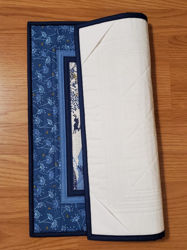 May include: A partially rolled-up quilted placemat with a dark blue border. The front features a blue floral pattern and a central panel with a rabbit illustration. The back is white with a quilted diamond pattern.