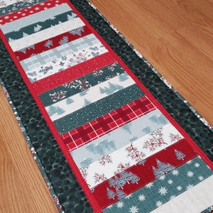 May include: A rectangular fabric table runner featuring a patchwork design of horizontal strips in red, white, and dark green. The patterns include stylized evergreen trees, snowflakes, plaid, and berry branches, all bordered by a dark green fabric.