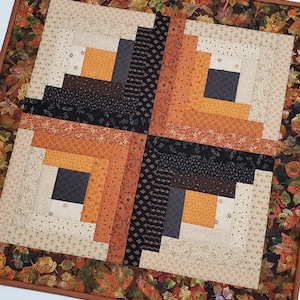 May include: A square fabric quilt featuring a log cabin block pattern in shades of black, orange, cream, and brown. The central design is surrounded by a border of fabric printed with a pattern of autumn leaves and finished with a brown binding.