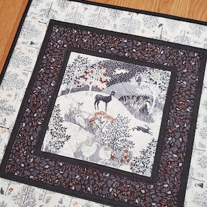 May include: A square quilted fabric mat featuring a central panel with a forest scene of a deer, birds, and rabbits in black, white, and orange. The center is framed by a dark charcoal border with a leaf pattern and an outer border of trees.
