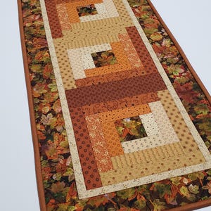 Puede incluir: Un camino de mesa rectangular con un patrón de colcha de cabaña de troncos en tonos naranja, marrón, crema y beige. El borde consiste en una tela oscura estampada con un patrón denso de hojas de otoño en tonos rojos, amarillos y marrones.