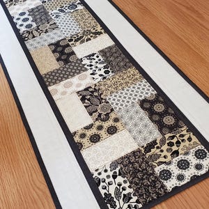 May include: A rectangular table runner featuring a patchwork design of various black, white, and beige fabric rectangles. The patterns include floral, geometric, and leaf motifs. The runner is bordered by a solid white strip and a thin black edge.