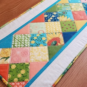 May include: A colorful quilted table runner with a patchwork design. The runner features squares of fabric in various patterns and colors, including sunflowers, floral prints, and geometric shapes. White and blue borders frame the patchwork.