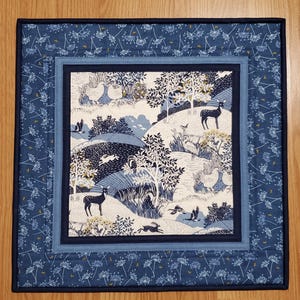 May include: A square quilted placemat with a nature scene. The center features a landscape with deer, birds, and trees in shades of blue, white, and yellow. The border has a floral pattern in navy blue and light blue.