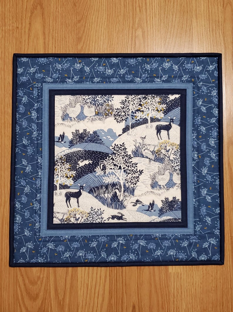 May include: A square quilted placemat with a nature scene. The center features a landscape with deer, birds, and trees in shades of blue, white, and yellow. The border has a floral pattern in navy blue and light blue.
