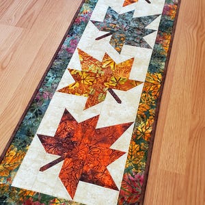May include: A rectangular quilted table runner featuring four large maple leaf appliques in shades of orange, red, and dark green. The center panel is cream-colored, surrounded by a border of multi-colored autumn leaves on a dark green background.