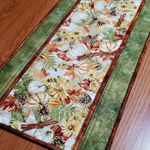 May include: A rectangular fabric table runner featuring a central panel with a print of white pumpkins, sunflowers, pinecones, cinnamon sticks, and autumn leaves on a white wood grain background, bordered by green and copper fabric.