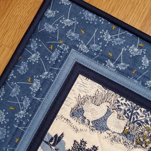 May include: A quilted placemat with a blue and white floral pattern. The placemat features a dark blue border and a central design with white chickens, trees, and a flying bird. The fabric has a quilted texture.