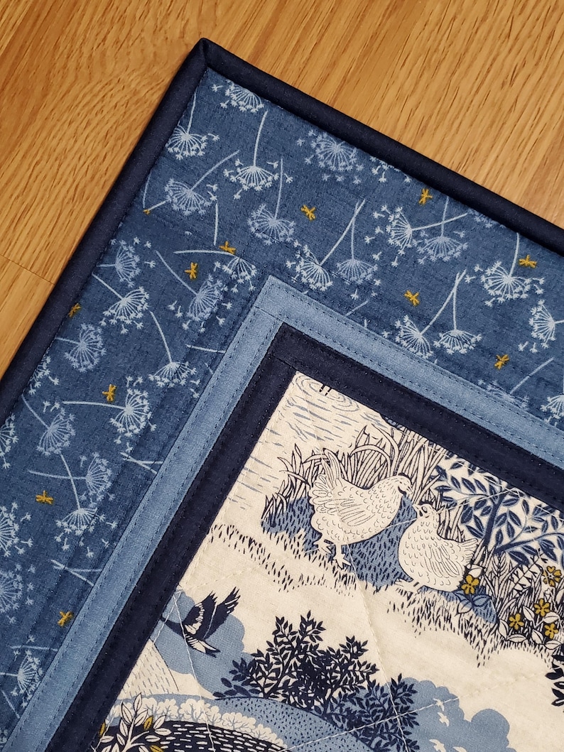 May include: A quilted placemat with a blue and white floral pattern. The placemat features a dark blue border and a central design with white chickens, trees, and a flying bird. The fabric has a quilted texture.