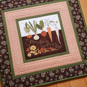 May include: A square quilted wall hanging featuring a central panel with illustrations of root vegetables and flowers. The text reads, "There is Peace in a Garden a quiet kind of Joy that only the Earth can give." It has borders in floral brown, solid green, and light pink fabric.