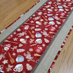 May include: A rectangular quilted table runner featuring a red background with a repeating pattern of gingerbread cookies, mugs of hot cocoa, wooden spoons, whisks, and the word joy. The runner has wide beige fabric borders on both sides.