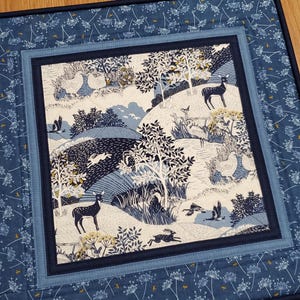 May include: A quilted placemat with a central panel depicting a woodland scene in shades of blue and white. The scene includes deer, rabbits, birds, and trees. The border is a darker blue with a floral pattern. The placemat is square.