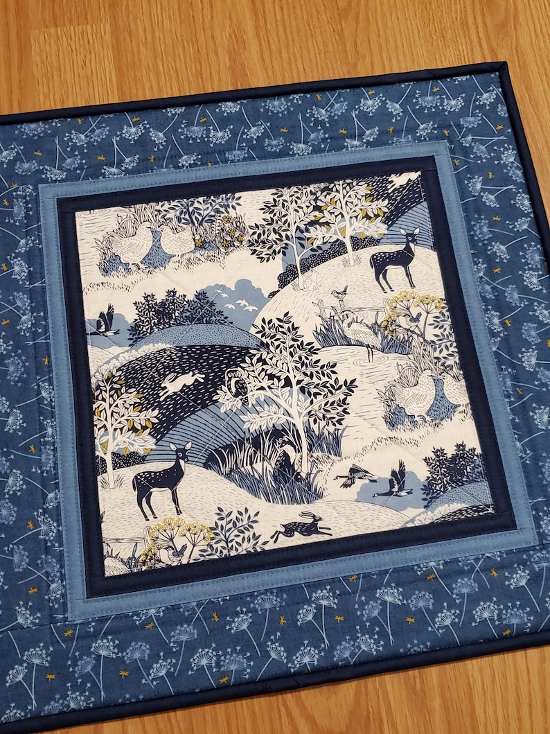 May include: A quilted placemat with a central panel depicting a woodland scene in shades of blue and white. The scene includes deer, rabbits, birds, and trees. The border is a darker blue with a floral pattern. The placemat is square.