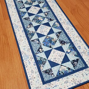 May include: A rectangular table runner with a patchwork design in shades of blue and white. The center features geometric shapes and floral patterns, framed by a border of small flowers and a solid blue edge. The runner is placed on a wooden surface.