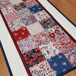 May include: A rectangular table runner featuring a patchwork design of various floral fabric squares in shades of red, blue, and white. The centre panel is bordered by a solid dark red strip and a white textured fabric, finished with a navy blue edge.