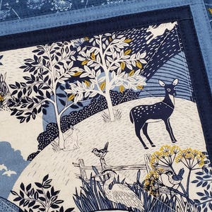 May include: A fabric panel with a nature scene featuring a deer, birds, and trees in shades of blue, white, and yellow. The panel has a dark blue border and a lighter blue patterned background.