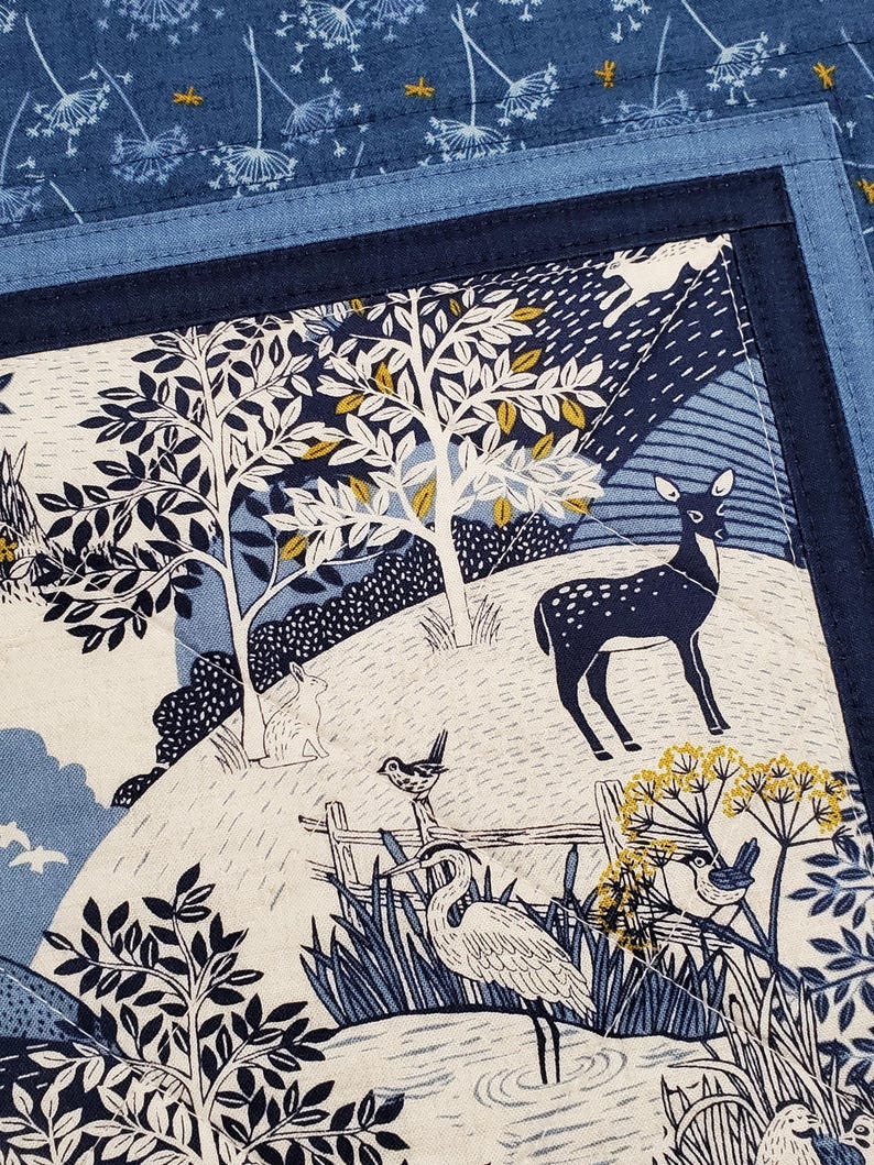 May include: A fabric panel with a nature scene featuring a deer, birds, and trees in shades of blue, white, and yellow. The panel has a dark blue border and a lighter blue patterned background.