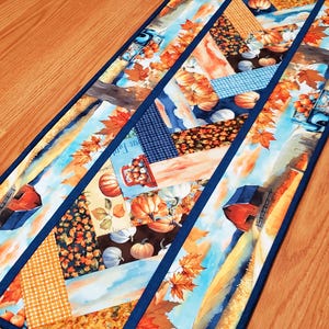 Può includere: Un runner da tavola rettangolare in tessuto con un design patchwork a tema autunnale che include zucche arancioni, fienili rossi, camion blu e fogliame autunnale nei toni dell'arancione, giallo e marrone, rifinito con un bordo blu scuro.