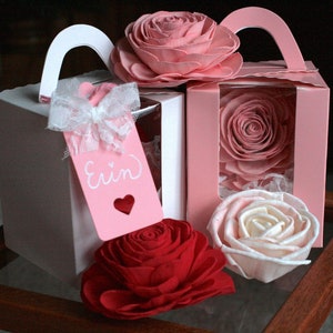 May include: Three pink and white paper boxes with a handle on top, each containing a single rose. The boxes are on a wooden table. One box has a pink tag with the name "Erin" written on it.