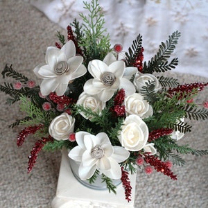 May include: A white and red Christmas floral arrangement in a metal bucket. The arrangement features white wooden flowers, red berries, and green foliage.
