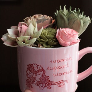 May include: Pink ceramic mug with a red graphic of a woman with flowers in her hair and the text "women support women". The mug is filled with a floral arrangement of pink and green succulents.