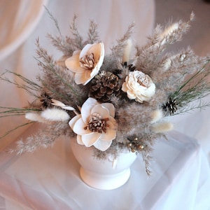 May include: A white ceramic vase holds a dried floral arrangement. The arrangement features beige pampas grass, white wooden flowers, pinecones, and sprigs of green. The flowers have brown centers. The background is a soft, white fabric.