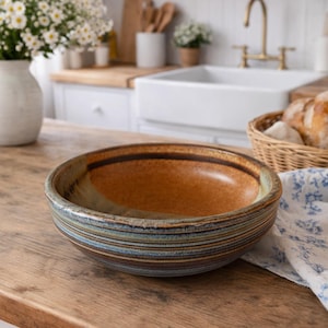 May include: A ceramic bowl with a brown interior and a striped exterior in shades of blue, brown, and tan. The bowl sits on a wooden surface in a kitchen setting. A basket of bread and a floral cloth are nearby.
