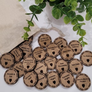 May include: Wooden discs with activity ideas like "Go Kart" and "Karaoke Night" are scattered. Each disc features a heart and a decorative border. They are spilling out of a beige drawstring bag, with text on each disc.