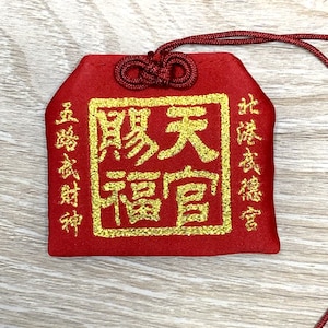 May include: A red fabric pouch with a gold embroidered square containing the Chinese characters "天照大神" (Ten-shoo Dai-jin) which translates to "The Great Shining Deity". The pouch has a red string attached to it.