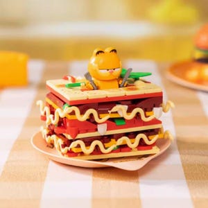 Op de afbeelding: Een speelgoedlasagne met een Garfield-figuur die een vork en mes vasthoudt. De lasagne is gemaakt van rode, gele en bruine blokken, met witte golvende noedels. Het staat op een klein, lichtgekleurd bord.
