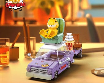 May include: A purple toy car with a Garfield character sitting in a green armchair on top. The car has a white roof and a sticker of Garfield on the side. The car is pulling a trailer with a popcorn container and a brown box.